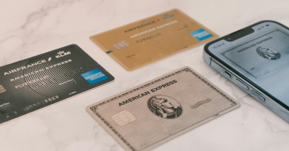 Three Amex credit cards plus one visible on a mobile phone screen sitting on a marble table.