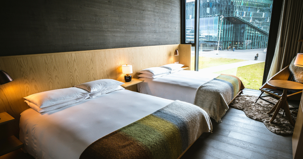 A hotel room with two queen beds that have a shared wooden headboard and fluffy striped blankets on the foot of each bed, with a city view visible through a floor to ceiling window, perfect for a military family who books through the Fedrooms program.