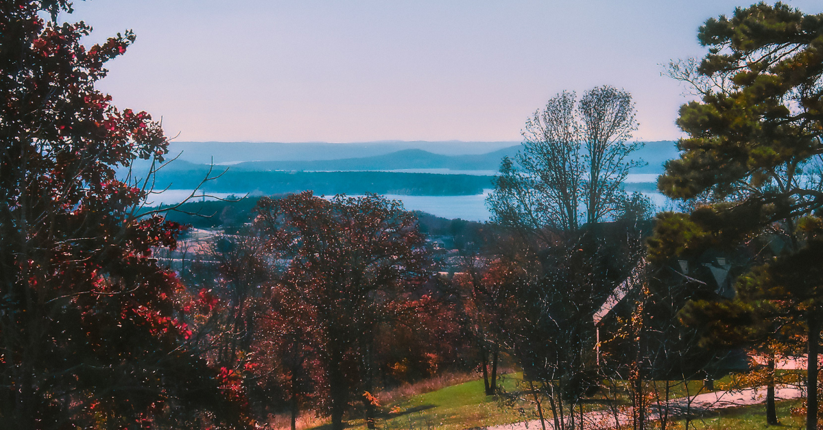 A view of the Ozarks near Ft Leonard Wood that shows a collection of red-leafed deciduous trees in the foreground and a lake visible in the background.
